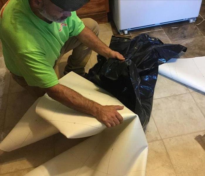 SERVPRO employee bent down on one knee, placing large pieces of flooring into a black bag 