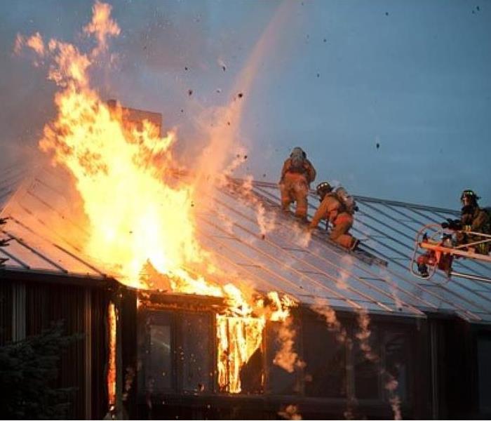 roof of a home up in flames with three firefighters on the roof with equipment 